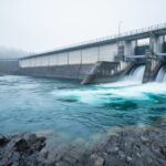 Hydroelectric dam and turbine intake with fast-flowing water on a rocky river under an overcast sky.