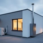 Modern house exterior at dusk showing an outdoor air-source heat pump unit and a conventional gas furnace vent on the building.
