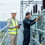 Two energy professionals inspecting electrical grid equipment at a modern substation with wind turbines and solar panels in the background.