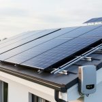Residential rooftop solar panels installed on a modern home, with sunlight reflecting off the photovoltaic glass and an inverter nearby.