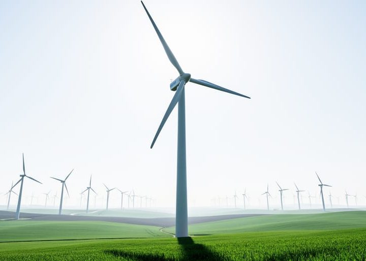 Modern wind turbines in a wind farm under an overcast sky, viewed from ground level with one turbine in sharp focus.