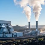 Geothermal power plant with steam rising from vent stacks and turbines under soft daylight, set against volcanic hills.