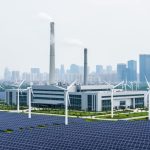 Modern industrial facility near a city skyline with faint vapor and nearby wind turbines and solar panels representing greenhouse-gas emissions reduction.