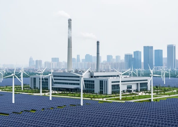 Modern industrial facility near a city skyline with faint vapor and nearby wind turbines and solar panels representing greenhouse-gas emissions reduction.