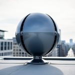 Spherical O-Wind turbine mounted on a rooftop under an overcast sky with city buildings blurred in the background.